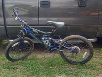 Side view of blue and black Excitor Magna dual suspension bike parked next to a truck, showing knobby tires, blue frame with black accents, padded seat, and rear dual suspension coil spring.