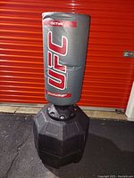 Full view of UFC OctTek punching bag with black weighted base, displayed outdoors on pavement before red door.