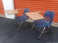 Two blue plastic chairs with attached wood tablet arms and metal wire baskets, placed outdoors on asphalt near an orange roll-up door.