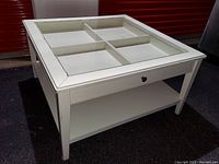 Front side angle of the white Ikea coffee table showing glass top divided into four panes, drawer with knob, and lower shelf.