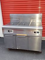 Front angle showing the stainless steel dual vat deep fryer unit with hinged lids, control knobs, and lower cabinet with two doors. Unit on casters.