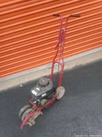 Full view of red metal frame yard edger with black Briggs & Stratton engine and handlebar, positioned on asphalt near orange door.