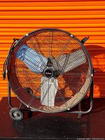 Front view of large utility fan showing three white blades in black metal protective cage with wheels at base.