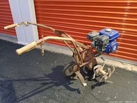 Full view of the tiller/cultivator showing wooden handles, metal frame, and blue engine mounted on top.