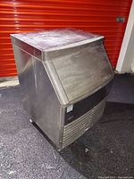 Front and side views of the stainless steel under counter ice machine showing rectangular shape, vented front panel, and access flap for ice.