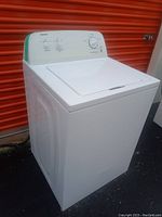 Front-right angled view of the white Admiral top-load washing machine set against an orange storage door background, showing the control panel with knobs and the closed lid.