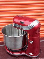 Front and side view of the red Dash Go stand mixer with stainless steel bowl attached on a dark surface, orange background in storage setting.