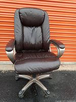 Front view of a black leather office chair with padding, armrests, and wheeled base against an orange storage door background.