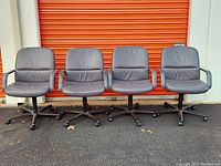 Four gray leather office chairs arranged side by side outdoors in front of an orange storage unit door. Chairs have cushioned seats and backs with armrests and rolling bases with casters.