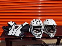 White and black STX lacrosse shoulder pad alongside two white lacrosse helmets with metal face cages placed on a wooden bench in front of orange metal door.