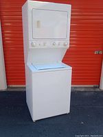 Front view of the stacked white laundry unit with washer and dryer outside against an orange door background.