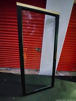 Glass storm door standing upright against a white and red background showing the full door with black frame and transparent glass.