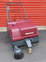 Front angle view of Maroon Minuteman commercial sweeper showing handlebar, large front body with logo, side brush, and caster front wheel.