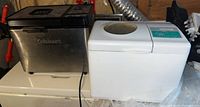 Two bread makers side by side on a surface; stainless steel Cuisinart and basic white Proctor Silex