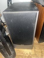Single black Quest speaker with cloth grille and black casing, positioned on a wooden floor near other furniture, showing minor dust and grime.
