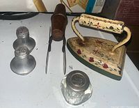 Photo showing all five items: antique cast iron clothing iron with floral painted design, two-blade cutter with wooden handle, pair of pewter salt and pepper shakers, and glass salt shaker with metal lid.