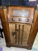 Front view of vintage wooden antique radio, showing control panel, knobs, and fabric speaker cover with some visible damage.