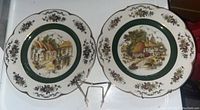 Two decorative wall plates with scalloped edges featuring detailed village scenes, floral borders, and gold trim, along with a metal plate stand on a white surface.