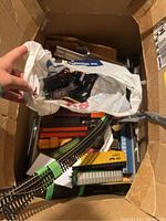 Box containing multiple curved metal tracks, train parts, and plastic bags with smaller items inside a cardboard box.