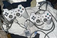 Two original Sony PlayStation controllers with wired connections shown lying on fabric background. Controllers have classic gray design with dual analog sticks and cable connectors visible.