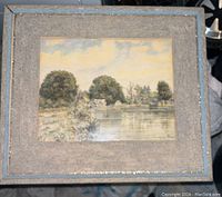View of the framed landscape painting with textured mat and blue frame, featuring house by water and trees under cloudy sky.
