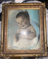Front view of framed baby portrait painting showing baby wrapped in white knitted blanket on blue background with artist signature.