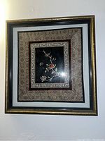 First framed fabric embroidered floral print showing a bird and various flowers on black fabric, surrounded by a patterned fabric mat and glass frame.