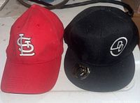 Red St. Louis Cardinals cap and black Dydi Loft cap placed side by side on a surface showing the front logos clearly.