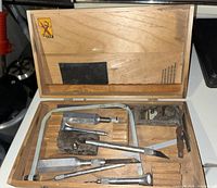 Open wooden case displaying various X-Acto tools including blades, knives with wooden handles, a small hand planer, and a metal hand saw with holder.