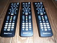 Three Shaw Direct remote controls placed on wood surface, showing front buttons and Shaw Direct branding.
