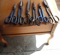 Photo shows six tin snips of varying sizes and handle types laid out on wooden furniture surface, blades and handles visible for condition assessment.