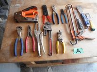 Photo showing a collection of 11 hand tools arranged on a wooden table with a green card marked 'A'. Tools include caulking gun, stapler, wire stripper, pliers, pipe wrench, hacksaw, scrapers.