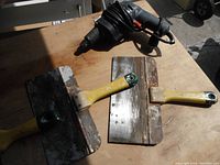 Photo showing two drywall trowels with yellow handles and rusty metal blades, next to a black corded electric drill.