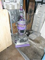 Dyson upright vacuum cleaner in gray and purple with wrapped power cord, standing on cement floor in storage room.