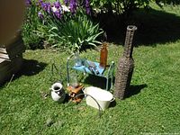 Photo showing decorative and garden items arranged on grass: a green garden kneeling stool, a tall wicker bottle-shaped vase, a white metal basin, a brown glass bottle, a white ceramic vase, black metal hooks, and a wasp catcher.