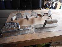 Seven used trowels including four brick trowels and three finishing trowels laid out on a wooden surface, showing different blade shapes and handle styles.