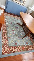 Rectangular floral patterned area rug with orange border in a room setting under a wooden table and near blue cabinet.