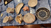 Assorted vintage ceramics including bowls, mugs, a plate, a pink bear salt shaker, and a shoelace hook laid on a blue cloth background