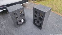 Pair of gray fabric covered rectangular box speakers with visible drivers and port holes, photographed outdoors on pavement.