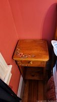Wooden bedside table shown in corner of room, displaying the floral painted details on the top and drawer, natural wood finish, and lower shelf.