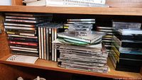 Photo of music CDs and cassette tapes stacked on a shelf showing a variety of cases and titles.