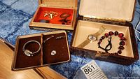 Three open jewelry boxes showing gold-tone wristwatch and red/black beaded necklace in one, small velvet case with silver watch and accessories, and additional orange-lined box with chain and earrings.