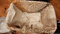 Overhead photo showing a variety of linens including a crocheted cream doily, an embroidered table runner, and several place mats with scalloped edges and brown stitching.