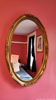 Front view of the oval wall mirror mounted on a red wall, showing reflection of a room with red walls, black curtains, framed artwork, and a bed.