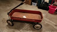 Full view of small red metal wagon with four wheels and black handle on a concrete floor.