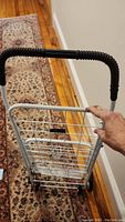 Top view of the collapsible metal shopping cart held upright showing black foam handle grips, white metal wireframe basket, and two wheels at bottom.