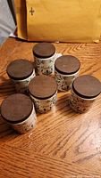Six ceramic spice canisters with wooden lids arranged on wooden surface, showing their tops and part of the decoration