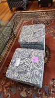 Photo showing two square storage ottomans with blue and white paisley fabric upholstery on a patterned rug next to a glass coffee table.