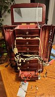 Upright wooden jewelry box opened showing mirror, drawers, side panels with hanging necklaces mostly in black, white, coral colors, and beaded styles, box is on a wood grain table.
