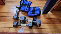 Photo showing three hex dumbbell iron hand weights (one 10 lb, two 5 lb) and two blue strap-on ankle weights on wood floor.